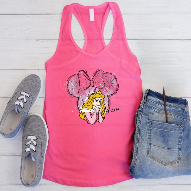 May include: A vibrant pink vest top with a Princess Aurora graphic. The design incorporates a Minnie Mouse-style bow and ears, with the name "Aurora" in script. Grey trainers and blue jeans are also in the image.