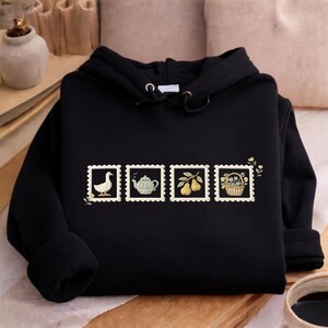 A black hoodie with four small illustrations on the front, including a duck, teapot, basket of fruit, and flowers, arranged in a row.