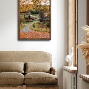 May include: A framed photograph of a country road winding through a field of autumn leaves, leading to a farmhouse with a red roof and a barn. The trees are in full fall foliage, with shades of yellow, orange, and red.