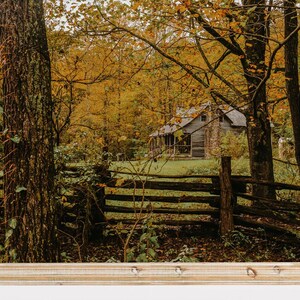 May include: A rustic wooden cabin nestled in a forest with autumn foliage. The cabin has a porch and a stone chimney. A wooden fence runs in front of the cabin, with trees and leaves in the foreground.