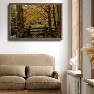 May include: A framed photograph of a rustic cabin in the woods. The cabin is surrounded by trees with yellow leaves, and a wooden fence runs in front of it.
