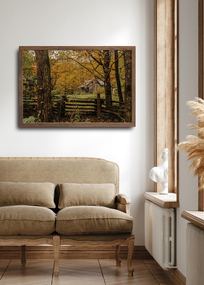 May include: A framed photograph of a rustic cabin in the woods. The cabin is surrounded by trees with yellow leaves, and a wooden fence runs in front of it.