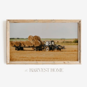 May include: A framed photograph of a blue tractor hauling a trailer full of hay bales in a field. The tractor is driving through a field of golden hay.