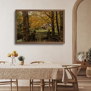 May include: A framed photograph of a rustic cabin nestled in a forest with autumn foliage. The cabin is surrounded by trees with yellow and orange leaves. A wooden fence runs in front of the cabin.