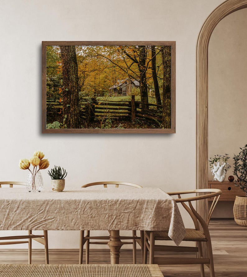 May include: A framed photograph of a rustic cabin nestled in a forest with autumn foliage. The cabin is surrounded by trees with yellow and orange leaves. A wooden fence runs in front of the cabin.