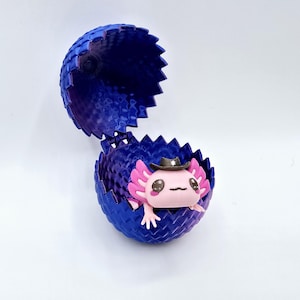 Puede incluir: Un ajolote rosa con un sombrero de vaquero negro se sienta dentro de un contenedor en forma de huevo azul y morado. El contenedor está abierto y el ajolote está mirando hacia afuera.