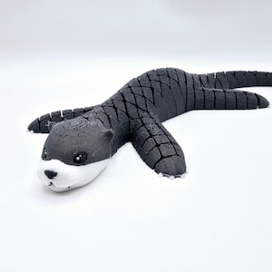 May include: A black and white 3D printed otter figurine with a textured, segmented body. The otter is lying on its belly with its head turned to the side.
