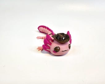 May include: A pink axolotl figurine wearing a small brown cowboy hat with a white star. The axolotl has pink gills and a pink tail. The figurine is on a white surface.