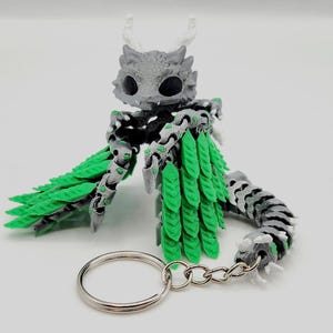 Puede incluir: Un llavero de dragón gris y verde con una cadena plateada. El dragón tiene una cola y alas de hojas verdes.