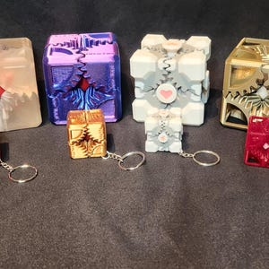 May include: A collection of 10 small, intricately designed, 3D printed cubes and spheres. The cubes are various colours, including blue, purple, gold, and white. The spheres are purple and black with intricate gear designs. Some of the cubes have keychains attached.