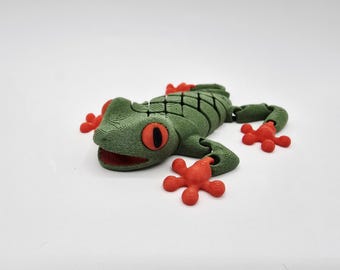 May include: A green and red frog figurine with a textured surface. The frog has large red eyes and red feet. The frog is lying on a white surface.
