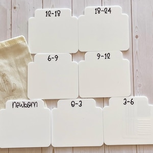 May include: White acrylic baby clothes size dividers with black text indicating sizes from newborn to 18-24 months. A beige fabric bag with a cactus illustration is on the left. The text "White acrylic with espresso paint" is at the bottom.