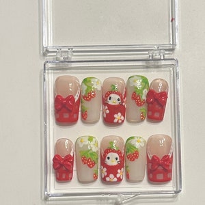 Może przedstawiać: Zestaw ośmiu sztucznych paznokci w stylu truskawkowo-Hello Kitty. Paznokcie są białe z czerwonymi i zielonymi akcentami. Każdy paznokieć ma czerwoną kokardkę i twarz Hello Kitty w czerwonej sukience z truskawką na głowie. Paznokcie są ułożone w przezroczystym plastikowym pudełku.