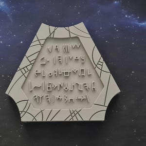 Puede incluir: Una placa geométrica blanca con un diseño intrincado y texto inscrito. La placa tiene la forma de un hexágono modificado, con una serie de líneas y formas que forman un borde. El texto parece estar en una escritura estilizada, no inglesa.