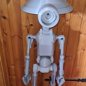 Op de afbeelding: Een grijs, 3D-geprint robotmodel met een groot, cirkelvormig oog en een conische kop. De robot heeft gelede ledematen en staat op een rode ondergrond tegen een houten achtergrond. Het heeft twee antennes.