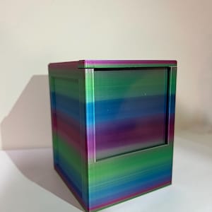 May include: A multi-colored, rectangular box with a sliding front panel. The box features a gradient of colors, including blue, green, purple, and pink. The box is made of a solid material and has a clean, modern design.