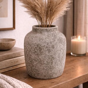 May include: A grey, textured vase filled with dried pampas grass. The vase sits on a wooden surface, with a candle in a glass and a framed picture in the background. The vase is approximately 25 cm tall.