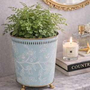 May include: A pale blue metal plant pot with a white floral pattern and gold trim, holding a green plant. The pot has small gold feet. A lit candle and perfume bottle are in the background.