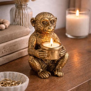 May include: A golden monkey-shaped candle holder with a lit candle. The monkey is sitting and holding a round candle holder. The candle holder is on a wooden surface, with a lit candle in the background.