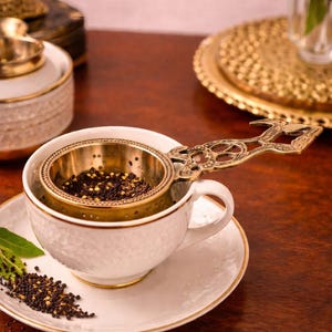Puede incluir: Una taza y un platillo blancos con ribete dorado, que sostienen un infusor de té de latón lleno de hojas de té. El infusor tiene un asa adornada. Las hojas de té están esparcidas sobre el platillo y la superficie de madera. Una bandeja dorada decorativa y flores están en el fondo.