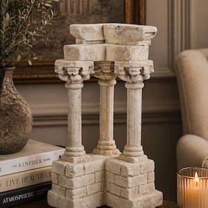 May include: A decorative, off-white stone architectural sculpture with three columns and a layered top. The sculpture is placed on a wooden surface, next to a vase with greenery, books, and a lit candle in a glass holder.