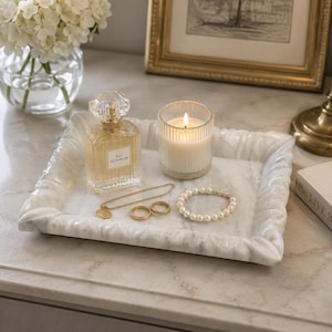 Puede incluir: Una bandeja rectangular de mármol blanco con borde festoneado que contiene un frasco de perfume de vidrio con la etiqueta Eau de Parfum, una vela de vidrio estriado con mecha encendida, una cadena dorada, dos anillos dorados y una pulsera de perlas.