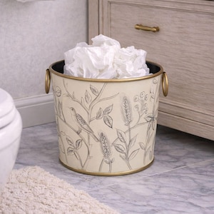 Puede incluir: Un cubo de basura decorativo de metal blanquecino con borde y asas doradas. El cubo presenta un diseño esbozado de un pájaro y plantas. El interior es negro y está lleno de pañuelos blancos. El cubo está en un baño.