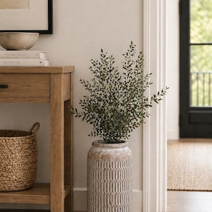 May include: A tall, textured, beige ceramic vase with a geometric pattern, filled with leafy green branches. The vase is placed near a wooden side table with a woven basket and books. The scene is set in a well-lit room with a neutral color palette.