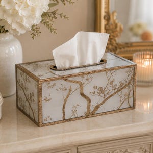 May include: A rectangular tissue box with a gold-colored frame and a white and gold floral design. The box is filled with white tissues and sits on a light-colored surface. A white vase with white flowers is in the background.
