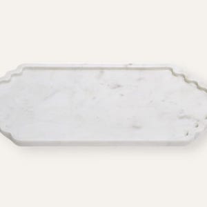 Op de afbeelding: Een rechthoekige witte marmeren dienblad met een geschulpte rand. De lade heeft een glad oppervlak met subtiele grijze adering. De algemene vorm is een lange rechthoek met decoratieve, golvende randen. Deze lade is geschikt om te serveren of tentoon te stellen.