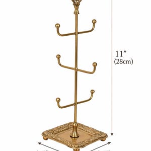 Pode incluir: Um suporte para canecas de metal dourado, com 28 cm de altura, com uma base quadrada de 9 cm de cada lado. A &aacute;rvore tem tr&ecirc;s n&iacute;veis de ganchos curvos para pendurar canecas e um topo decorativo.