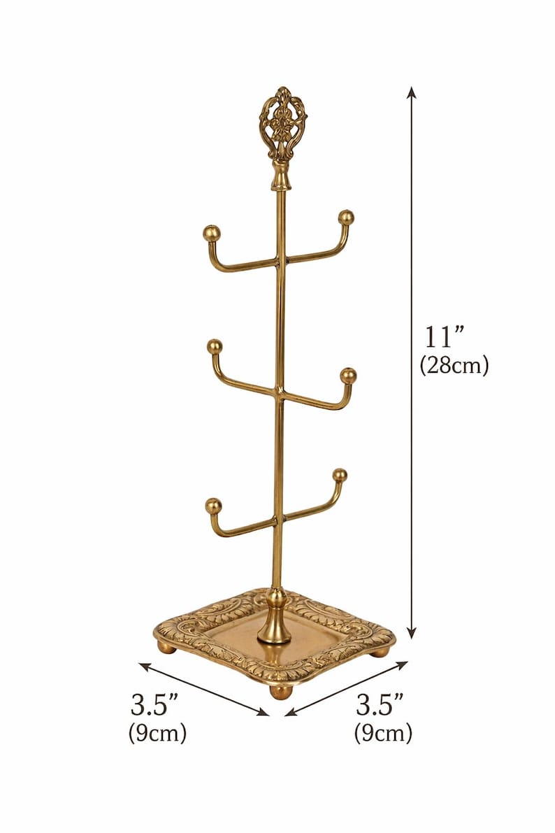 Pode incluir: Um suporte para canecas de metal dourado, com 28 cm de altura, com uma base quadrada de 9 cm de cada lado. A &aacute;rvore tem tr&ecirc;s n&iacute;veis de ganchos curvos para pendurar canecas e um topo decorativo.