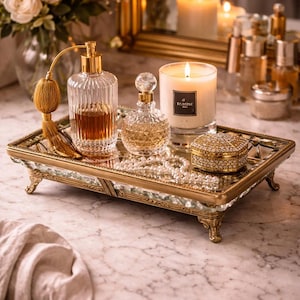 May include: A gold-toned vanity tray displaying perfume bottles, a candle, and a bejeweled box. The tray has intricate detailing and holds a lit candle and several perfume bottles. A bouquet of white flowers and a mirror are visible in the background.