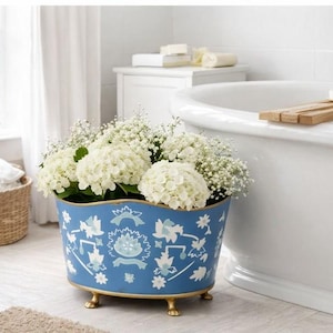 May include: A blue oval planter with white floral patterns and gold feet, filled with white flowers. The planter is in a bathroom setting, near a white bathtub and a window with white curtains. A woven basket is visible.