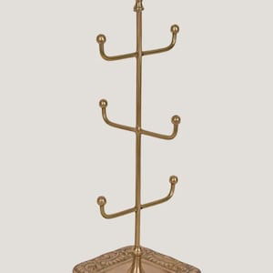Pode incluir: Uma &aacute;rvore de canecas de metal dourado com base e topo decorativos. A &aacute;rvore tem quatro bra&ccedil;os para segurar canecas, e a base &eacute; quadrada com detalhes ornamentados. O topo da &aacute;rvore tem um remate decorativo.