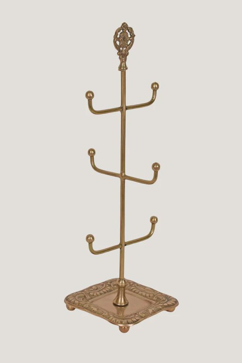 Pode incluir: Uma &aacute;rvore de canecas de metal dourado com base e topo decorativos. A &aacute;rvore tem quatro bra&ccedil;os para segurar canecas, e a base &eacute; quadrada com detalhes ornamentados. O topo da &aacute;rvore tem um remate decorativo.