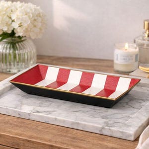 May include: A rectangular black tray with red and white striped interior, gold trim, and a marble base. The tray is displayed with a vase of white flowers, a candle, perfume, and a starfish.