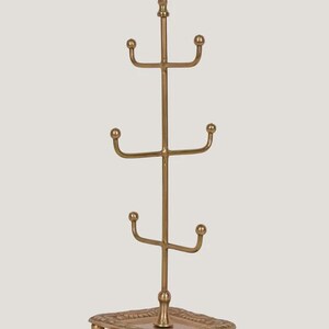 Pode incluir: Um suporte para canecas de metal dourado com base quadrada e topo decorativo. A &aacute;rvore tem quatro n&iacute;veis de ganchos curvos para pendurar canecas. O topo da &aacute;rvore tem um design ornamentado.