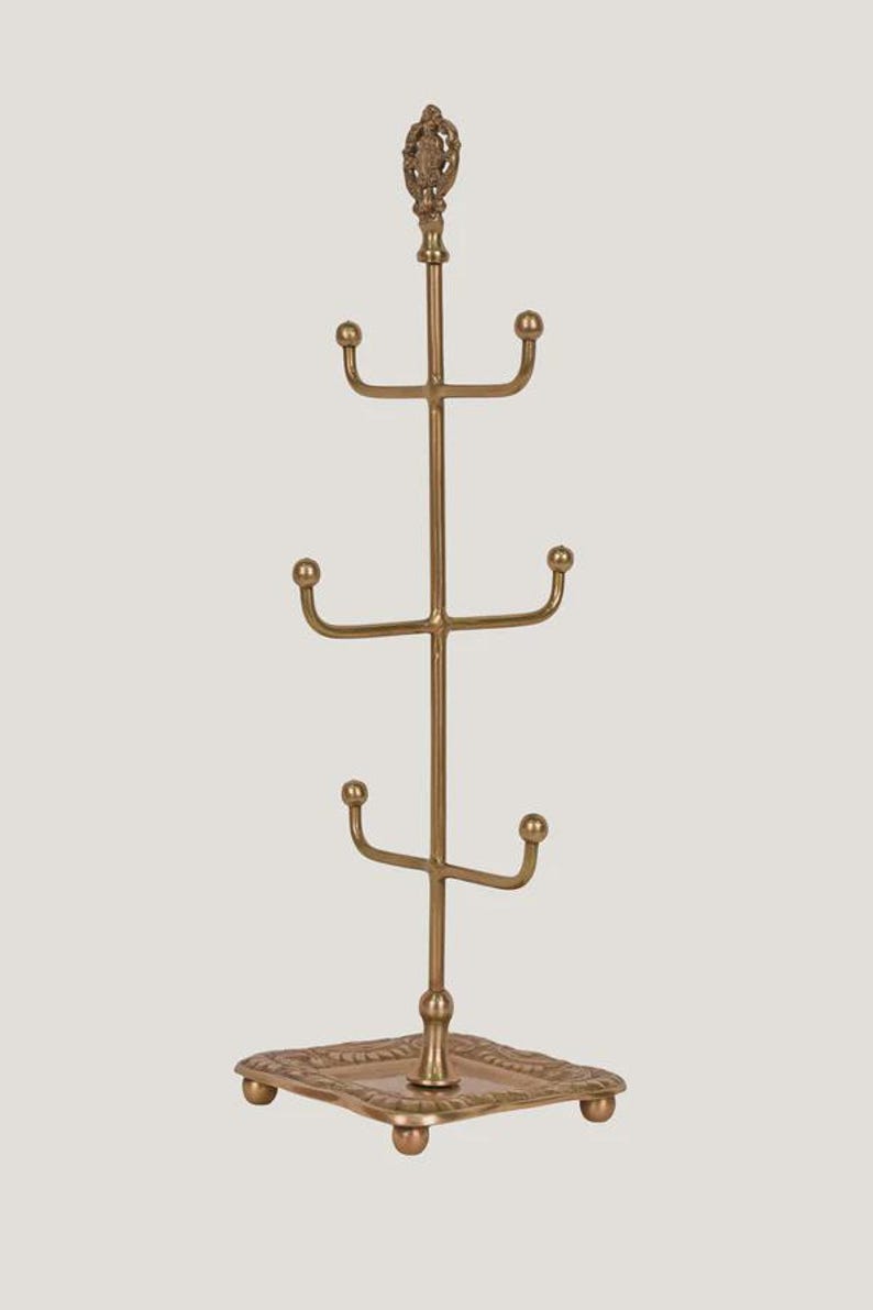 Pode incluir: Um suporte para canecas de metal dourado com base quadrada e topo decorativo. A &aacute;rvore tem quatro n&iacute;veis de ganchos curvos para pendurar canecas. O topo da &aacute;rvore tem um design ornamentado.