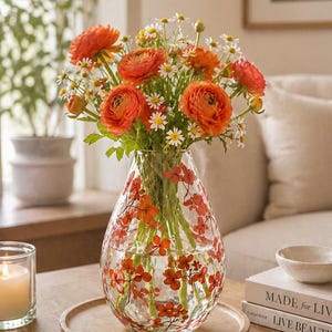 Op de afbeelding: Een heldere glazen vaas met oranje bloemmotieven bevat een boeket van oranje ranonkels en witte madeliefjes. De vaas staat op een houten dienblad, met een brandende kaars en boeken in de buurt. De bloemen zijn het middelpunt.