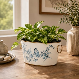 May include: An oval white metal planter featuring a blue painted design of a bird perched on a flowering branch. The planter has two gold-toned ring handles on the sides and contains a green leafy plant with variegated yellow and green foliage.