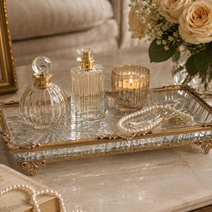 May include: A decorative glass tray with gold trim holds perfume bottles, a lit candle, and jewelry. The tray sits on a marble surface, with a book and a pearl necklace in the foreground. A vase of roses is in the background.