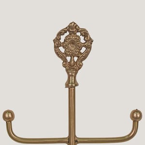 Pode incluir: Um suporte de joias de metal dourado com um topo ornamentado e decorativo. O suporte tem um poste central vertical com dois bra&ccedil;os curvos que se estendem horizontalmente, cada um terminando em uma pequena bola. O topo apresenta um design floral detalhado.