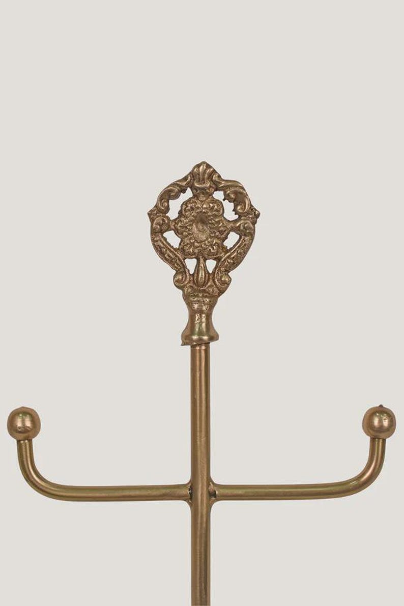 Pode incluir: Um suporte de joias de metal dourado com um topo ornamentado e decorativo. O suporte tem um poste central vertical com dois bra&ccedil;os curvos que se estendem horizontalmente, cada um terminando em uma pequena bola. O topo apresenta um design floral detalhado.