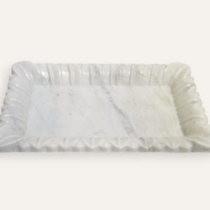 Puede incluir: Una bandeja rectangular de mármol blanco con un borde decorativo y acanalado. La superficie de la bandeja tiene sutiles vetas grises, lo que se suma a su apariencia de piedra natural. Las esquinas presentan un diseño estilizado y ondulado.