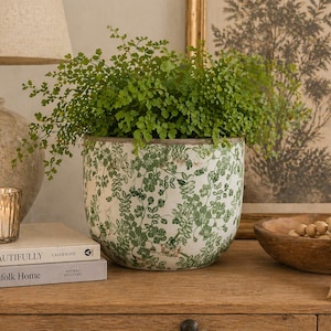 Op de afbeelding: Een witte keramische plantenpot met een groen bladerpatroon, gevuld met een weelderige groene plant. De pot staat op een houten oppervlak, naast boeken, een kaars en een decoratieve kom. De algehele esthetiek is een mix van natuurlijke en decoratieve elementen.