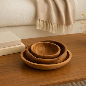 Puede incluir: Un juego de tres cuencos de madera de diferentes tamaños, anidados entre sí. Los cuencos son de madera de color marrón claro y tienen un acabado liso y natural. El cuenco más grande tiene unos 25 cm de diámetro.