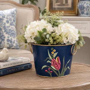 Pode incluir: Um vaso de metal em azul-marinho profundo com um design pintado de uma tulipa rosa com folhas verdes e pequenas flores amarelas. O balde tem uma borda dourada e duas alças circulares douradas nas laterais, preenchido com flores de hortênsia brancas.