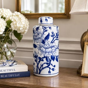 May include: A tall, cylindrical ceramic jar with a lid, featuring a blue and white floral design. The jar is decorated with large peony blooms and delicate branches. It is placed on a wooden surface, alongside a lamp and a framed picture.