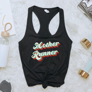 K&ouml;nnte beinhalten: Schwarzes Tanktop mit dem Text "Mother Runner" in einer Retro-Schriftart mit gr&uuml;nen, roten und gelben Farben. Das Tanktop liegt auf einer wei&szlig;en, flauschigen Oberfl&auml;che.